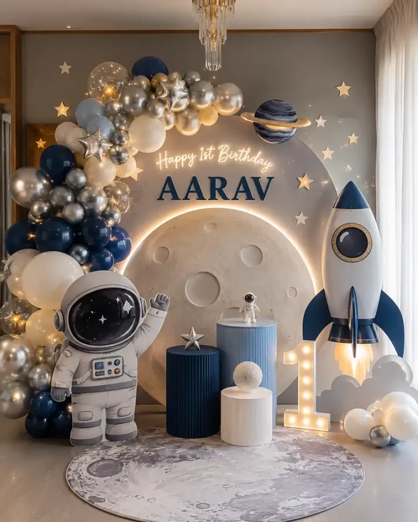 Space-themed first birthday backdrop featuring a large illuminated moon, blue, silver, and white balloon garland, astronaut cutout, rocket ship decoration, planets and stars, glowing number one marquee, and personalized “Happy 1st Birthday Aarav” sign in an elegant indoor celebration setup