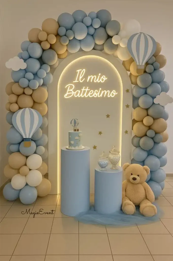 Blue and cream baptism celebration backdrop with a balloon arch, illuminated arched panel reading “Il mio Battesimo,” hot-air-balloon decorations, teddy bear, pedestal stands, and a small themed cake in a soft pastel color palette.