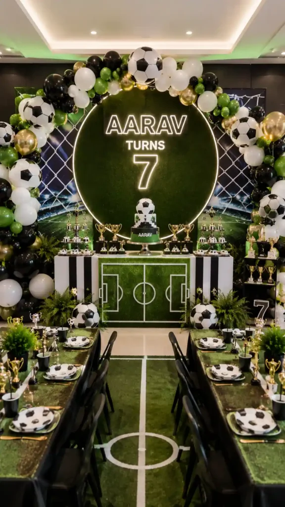 Soccer-themed birthday party setup featuring a green turf-inspired backdrop, soccer ball balloon garlands, and a glowing sign reading 'Aarav Turns 7.' The celebration includes a soccer-themed cake, trophy displays, football field graphics, black, white, green, and gold decorations, and long dining tables styled like a soccer pitch, creating an exciting sports-themed party atmosphere.
