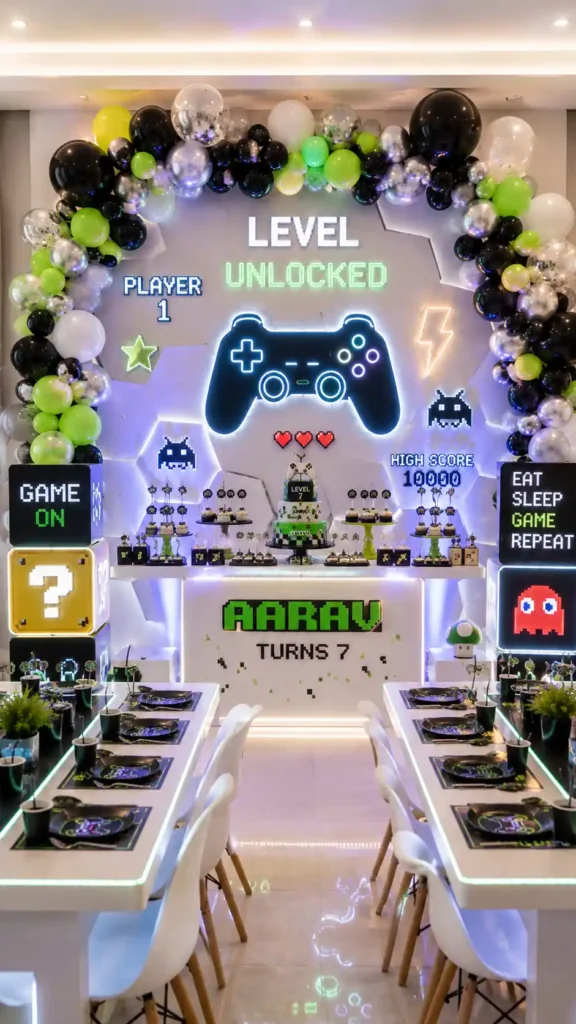 Modern video game–themed birthday party setup featuring neon-inspired décor, a glowing game controller centerpiece, and pixel-art graphics. The backdrop displays 'Level Unlocked' and 'Aarav Turns 7,' surrounded by black, white, silver, and lime-green balloon garlands. Gaming icons, arcade-style elements, themed desserts, illuminated tables, and interactive visual effects create an exciting gamer celebration atmosphere.