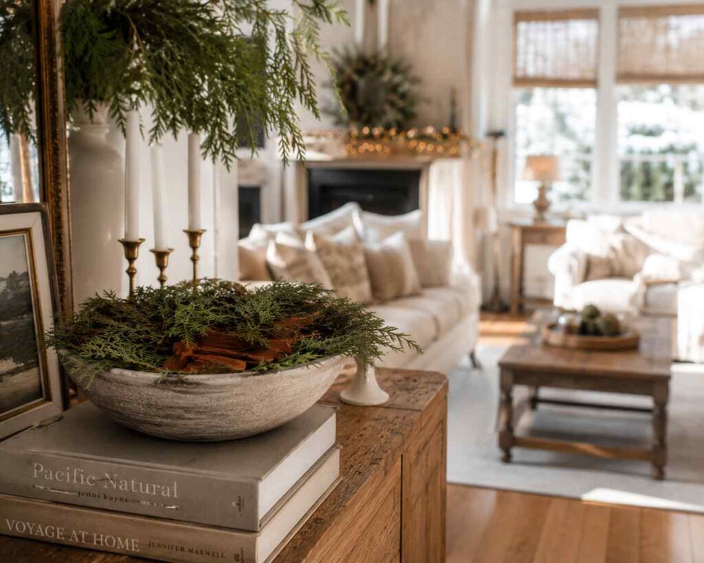 Close-up of a rustic wooden console table decorated for Christmas, featuring a textured bowl filled with evergreen branches and cinnamon sticks resting on stacked coffee-table books. In the softly blurred background, a bright living room with cream sofas, a coffee table, garland lights, and seasonal décor creates a warm and inviting holiday atmosphere