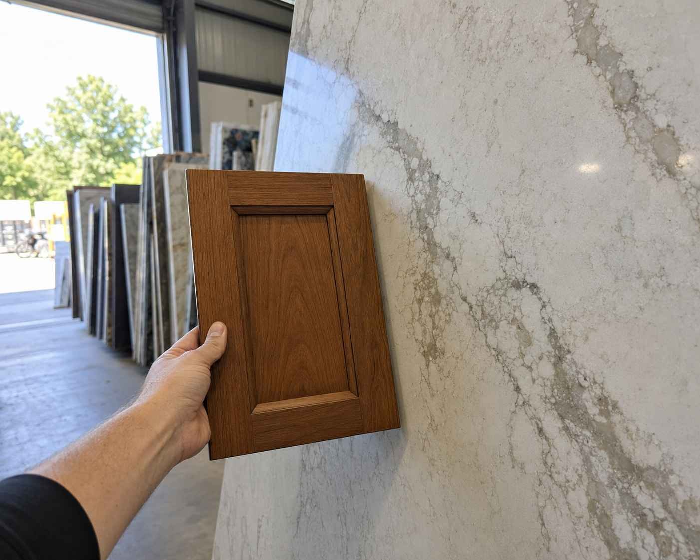 Hand holding a walnut wood cabinet door sample against a large veined quartz slab in a stone yard, illuminated by natural daylight with rows of stone slabs blurred in the background for a realistic kitchen material comparison.