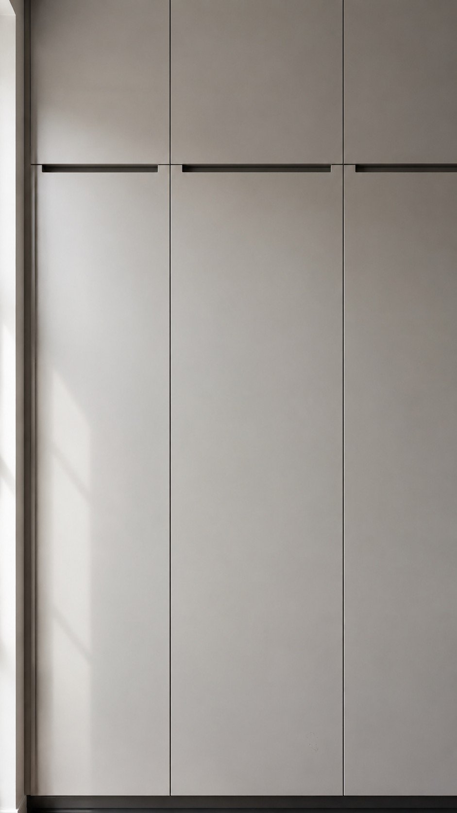 Floor-to-ceiling light grey slab kitchen cabinets with integrated handleless pulls, smooth satin finish, matte black base trim, and soft natural daylight in a modern minimalist interior.