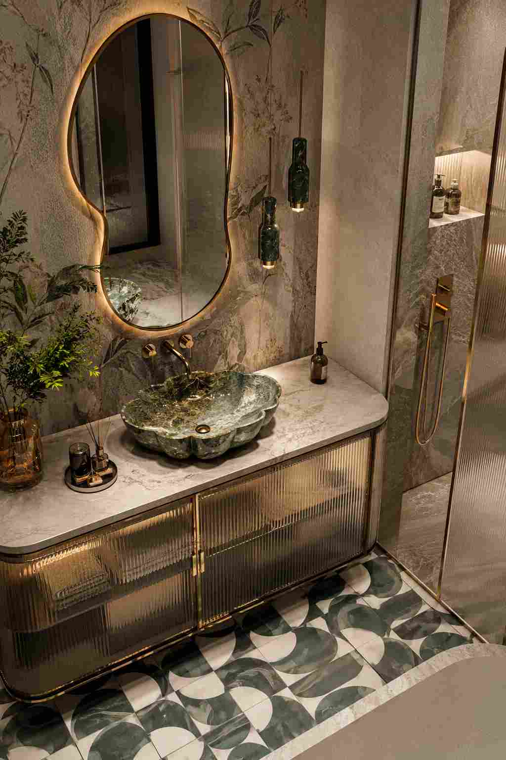 elegant luxury bathroom featuring a sculptural green marble vessel sink on a stone vanity with fluted glass cabinets, an organically shaped backlit mirror, brass fixtures, floral-patterned wall tiles, a walk-in shower, and geometric green-and-white floor tiles.