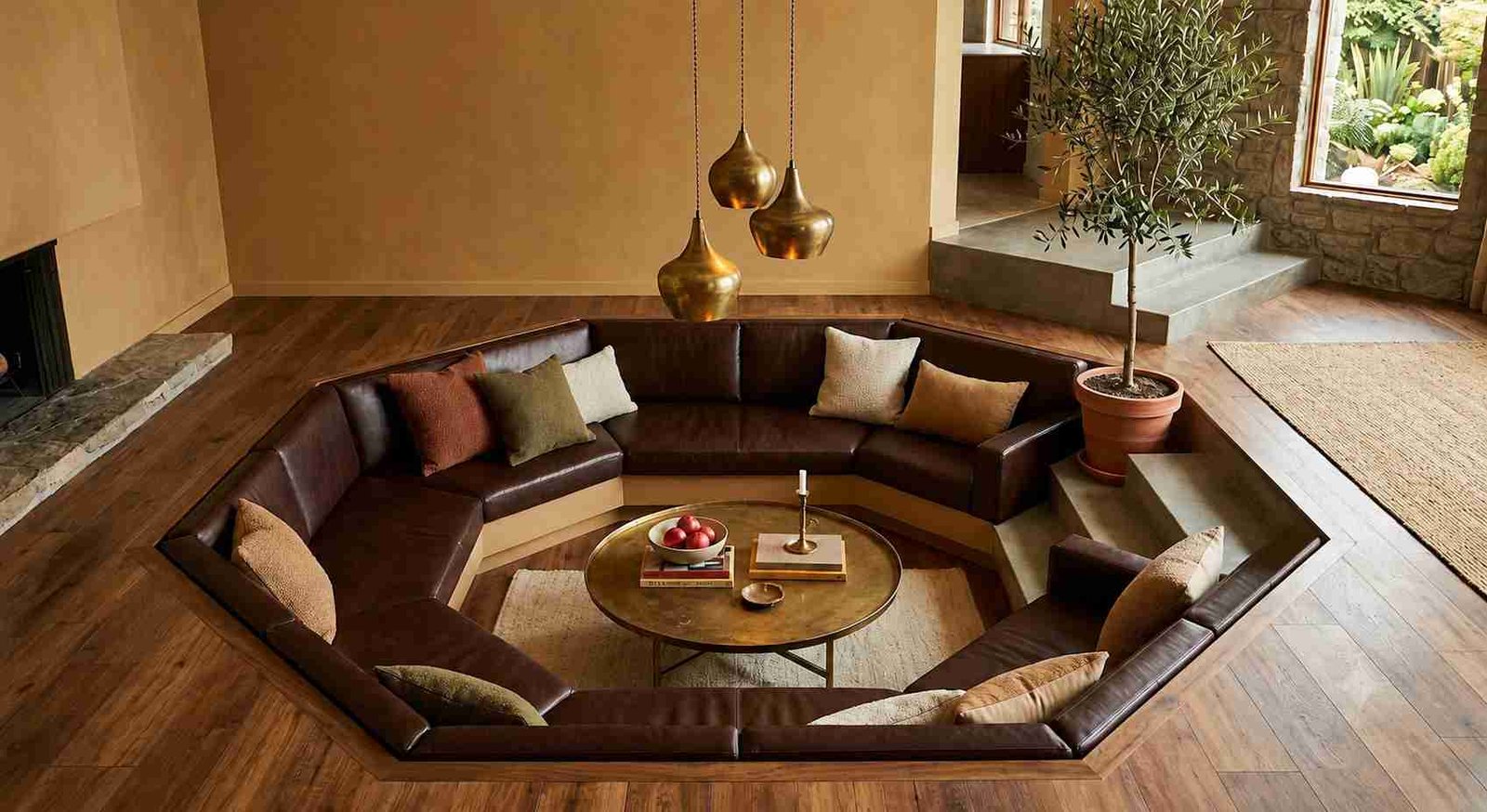 Sunken conversation pit living room with chocolate brown leather sectional, stone table, brass lamp, and a real olive tree by the window.